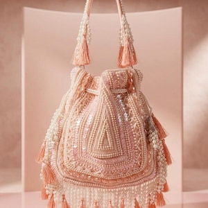 Puede incluir: Un bolso rosa rubor con cierre de cordón, adornado con perlas, lentejuelas y borlas. El bolso presenta un asa con cuentas y está adornado con intrincados diseños. Las borlas son de color melocotón suave.