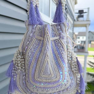 May include: A lavender beaded handbag with white pearl accents and matching tassels. The bag features a drawstring closure and a decorative pattern of beads and sequins. The handle is also adorned with pearls and tassels, creating an elegant accessory.