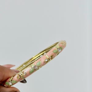 K&ouml;nnte beinhalten: Ein goldfarbener Armreif mit einer Basis aus zartrosa Emaille, verziert mit hellgr&uuml;nen Blattmotiven und kleinen, klaren Kristallen, die in die Mitte jedes Blumendesigns eingelassen sind und ein feines, wiederkehrendes Muster bilden.