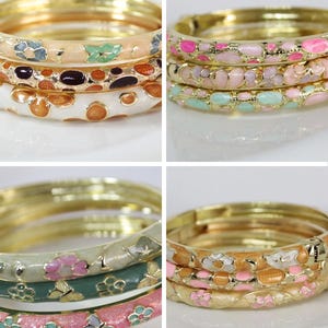Puede incluir: Cuatro conjuntos de brazaletes de metal en tono dorado, cada uno con tres pulseras. El set 1 presenta patrones florales y abstractos en crema, naranja y marrón. El set 2 tiene incrustaciones ovaladas en rosa, lavanda y verde menta. El set 3 muestra motivos florales y de mariposas en salvia, rosa y crema. El set 4 exhibe flores rosas y blancas sobre aros dorados.
