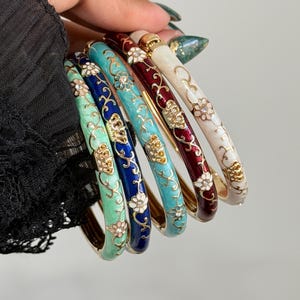 May include: A set of five hinged bangle bracelets in mint green, royal blue, turquoise, deep red, and cream. Each metal bracelet features a gold-tone floral vine pattern with small clear crystals embedded in the center of the flower motifs.