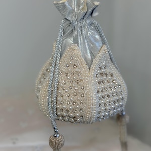 Puede incluir: Un bolso con cordón de cuentas plateadas y blancas con un diseño floral. El bolso presenta una parte superior metálica plateada, cuentas de perlas y brillantes pedrería. Tiene asas de cuerda plateada y borlas de cuentas.