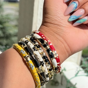 Op de afbeelding: Een verzameling kleurrijke emaille armbanden gestapeld op een pols. De armbanden hebben bloemen- en geometrische ontwerpen in geel, zwart, wit en rood, geaccentueerd met goud en strass. De nagels zijn geverfd met een lichtroze basis en blauwe en groene wervelingen.