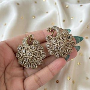 May include: A pair of gold-coloured peacock earrings. Each earring has a peacock design with a head and a fan-shaped tail. The earrings are encrusted with clear gemstones and a small red gemstone on the head.