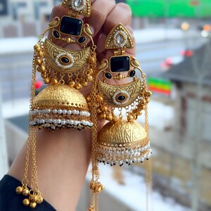 May include: A pair of gold-toned earrings with intricate detailing. Each earring features a triangular top, a black rectangular stone, and a bell-shaped bottom. White and black beads and dangling chains add to the design. The earrings are held up.