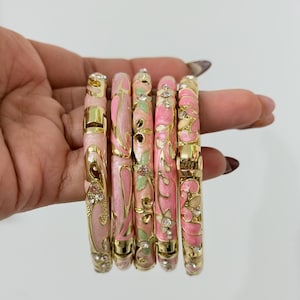 K&ouml;nnte beinhalten: Ein Set aus f&uuml;nf aufklappbaren Armreifen mit goldfarbenen Metallrahmen, verziert mit rosa und hellgr&uuml;nen Emaille-Blumen- und Wirbelmustern sowie kleinen, in die Oberfl&auml;che eingelassenen klaren Strasssteinen.