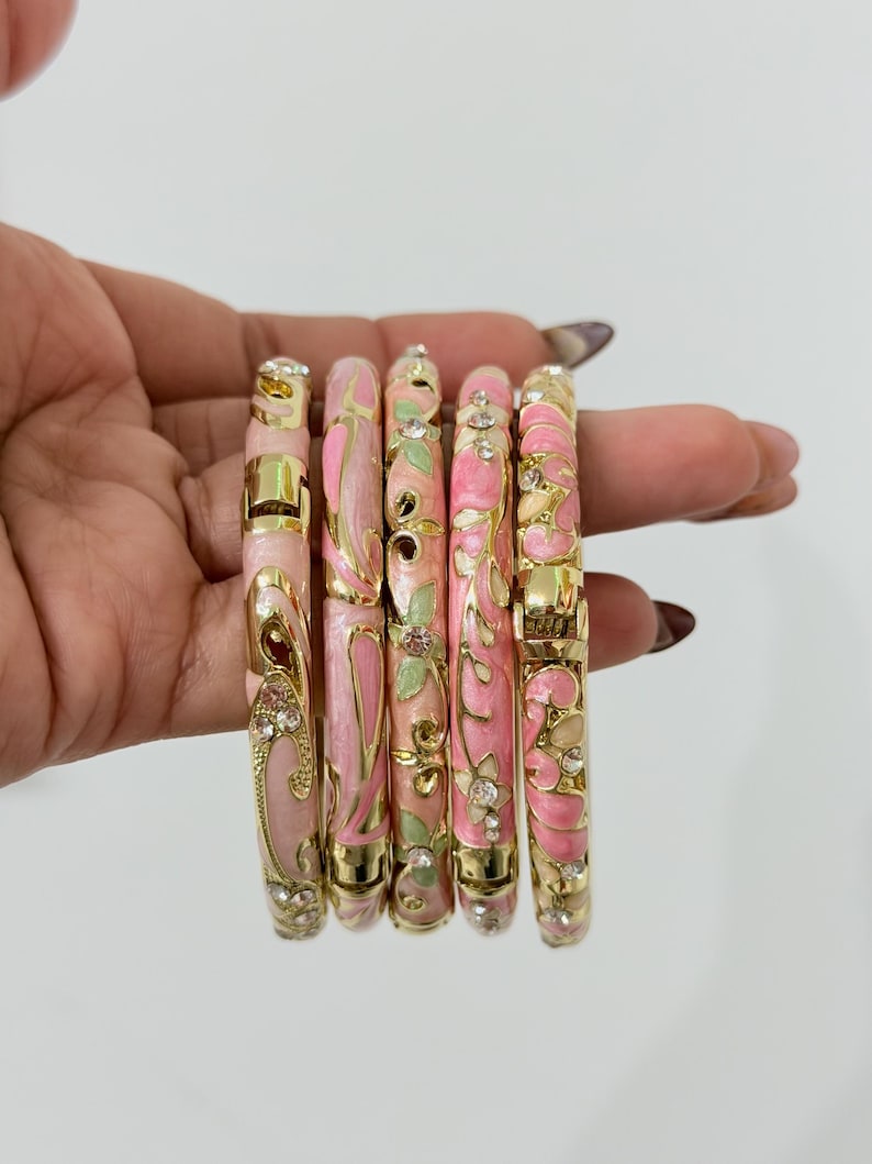K&ouml;nnte beinhalten: Ein Set aus f&uuml;nf aufklappbaren Armreifen mit goldfarbenen Metallrahmen, verziert mit rosa und hellgr&uuml;nen Emaille-Blumen- und Wirbelmustern sowie kleinen, in die Oberfl&auml;che eingelassenen klaren Strasssteinen.
