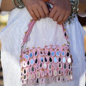 Könnte beinhalten: Eine rosa Handtasche mit einem dekorativen Muster aus tropfenförmigen Verzierungen. Die Tasche hat einen passenden rosa Riemen und ist mit goldfarbenen Akzenten verziert. Die Tasche wird von einer Person gehalten, die ein weißes Kleid und silberne Armbänder trägt.