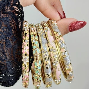 Könnte beinhalten: Eine Sammlung goldener Armreifen mit Blumen- und Blattmotiven in Rosa, Weiß und Mintgrün. Jeder Armreif weist aufwendige Details auf und ist mit kleinen, funkelnden Kristallen verziert. Die Armreifen werden von einer Hand mit rotem Nagellack gehalten.