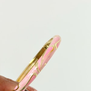 K&ouml;nnte beinhalten: Ein aufklappbarer Armreif mit einer Basis aus poliertem goldfarbenem Metall und einer Einlage aus wirbelndem rosa Emaille. Das Design zeigt ein durchgehendes Muster aus rosa Emaille-Abschnitten, die durch d&uuml;nne, erhabene goldfarbene Metalllinien getrennt sind.