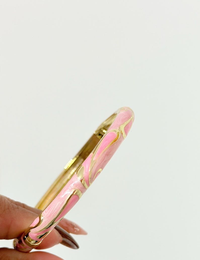K&ouml;nnte beinhalten: Ein aufklappbarer Armreif mit einer Basis aus poliertem goldfarbenem Metall und einer Einlage aus wirbelndem rosa Emaille. Das Design zeigt ein durchgehendes Muster aus rosa Emaille-Abschnitten, die durch d&uuml;nne, erhabene goldfarbene Metalllinien getrennt sind.
