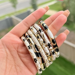 May include: A collection of seven white, black, and gold bangles. The bangles feature floral and geometric designs. Some bangles have black floral patterns, while others have white backgrounds with black accents. The bangles are held in a hand.