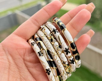 May include: A collection of seven white, black, and gold bangles. The bangles feature floral and geometric designs. Some bangles have black floral patterns, while others have white backgrounds with black accents. The bangles are held in a hand.
