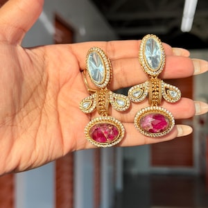Peut inclure: Une paire de boucles d'oreilles pendantes dorées avec un cristal ovale transparent en haut, un élément central en or texturé et un pendentif ovale en bas contenant des pétales de fleurs roses incrustés, bordés de petites pierres transparentes.