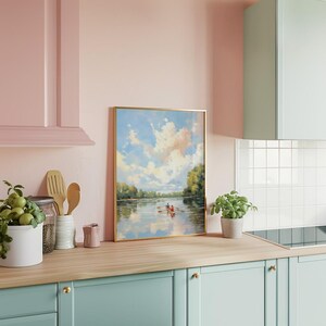 May include: A framed landscape painting with a gold frame depicts a river scene with two people in a rowboat under a cloudy sky. The painting is displayed on a light wood countertop in a kitchen with pink and blue cabinets.