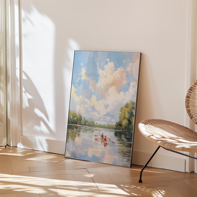 May include: A framed landscape painting depicts a serene river scene under a cloudy sky. The artwork features a boat with two figures rowing on the water, surrounded by lush green trees along the riverbanks. The painting is leaning against a white wall.