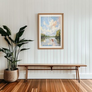 May include: A framed landscape painting depicts a river scene with a boat and figures under a cloudy sky. Below, a long wooden bench with a woven seat sits on a hardwood floor. A large potted plant is on the left, against a white paneled wall.