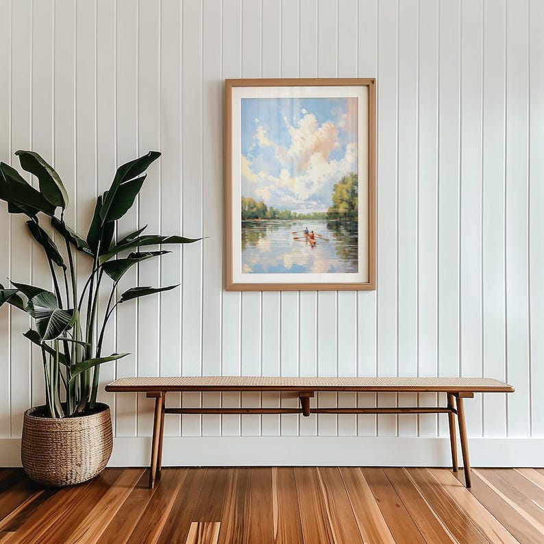 May include: A framed landscape painting depicts a river scene with a boat and figures under a cloudy sky. Below, a long wooden bench with a woven seat sits on a hardwood floor. A large potted plant is on the left, against a white paneled wall.