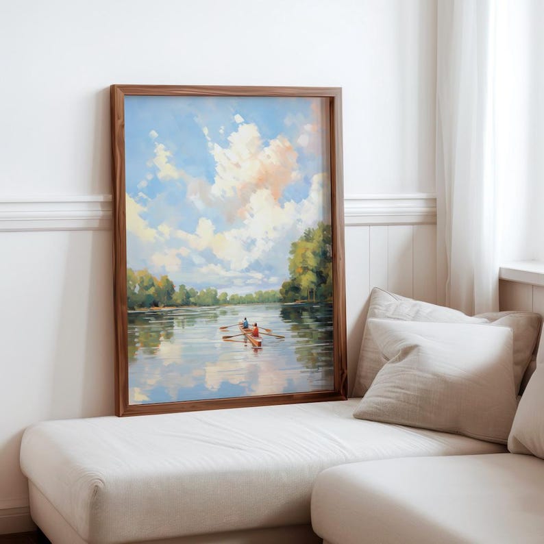 May include: A framed landscape painting depicts a serene river scene with a rowboat. The artwork features a blue sky with fluffy clouds, lush green trees, and the reflection of the scenery in the water. The frame is made of dark wood.
