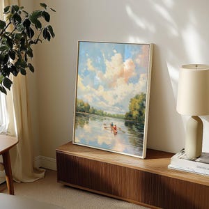 May include: A framed landscape painting depicts a serene river scene with a boat and two figures rowing. The sky is filled with fluffy clouds, and the water reflects the sky and surrounding trees. The painting is displayed on a wooden cabinet.