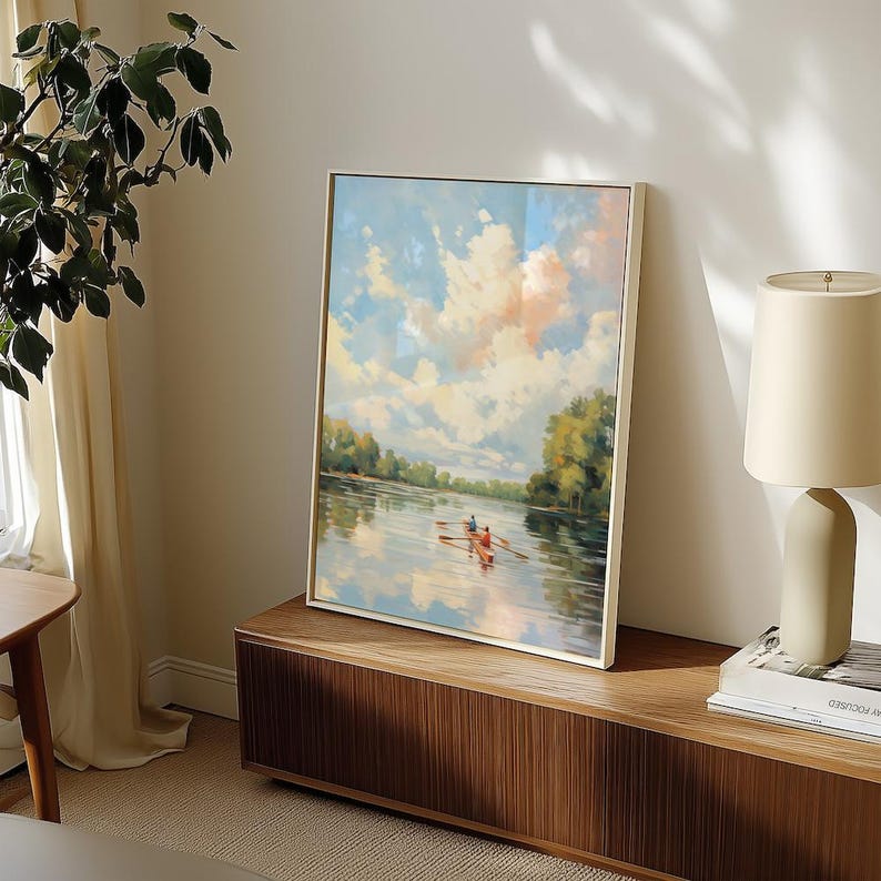 May include: A framed landscape painting depicts a serene river scene with a boat and two figures rowing. The sky is filled with fluffy clouds, and the water reflects the sky and surrounding trees. The painting is displayed on a wooden cabinet.