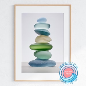 May include: A framed print featuring a stack of smooth, colorful sea glass stones. The stones are in shades of blue, green, and cream, balanced on top of each other. The print is in a wooden frame and has a white border.