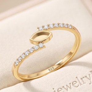 May include: A gold-toned ring featuring a marquise-shaped setting in the center, flanked by two rows of small clear stones along the band. The interior of the band is stamped with the number 925, indicating sterling silver composition.