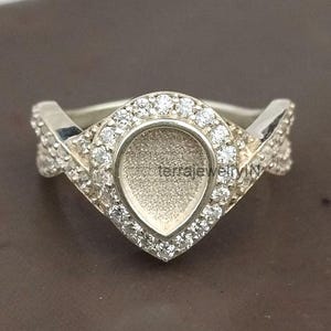 May include: A silver ring with a teardrop-shaped center setting, surrounded by small, clear gemstones. The band features a V-shaped design with additional gemstones. The ring is displayed on a brown surface.