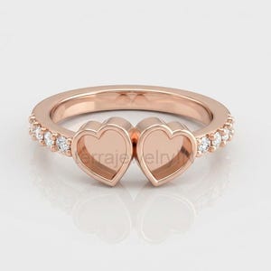 May include: Rose gold ring with two heart-shaped settings. The band is set with small, sparkling white gemstones. The ring is displayed on a white surface, highlighting its reflective qualities. A romantic piece of jewellery.