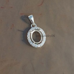 May include: Silver oval pendant with a halo of small, clear stones around the central setting. The pendant has a small loop at the top for a chain. The text "terra jewelryIN" is visible on the side.