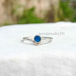 May include: A silver ring with a blue gemstone set in a V-shaped band. The ring is on a white surface. terrajewelryIN