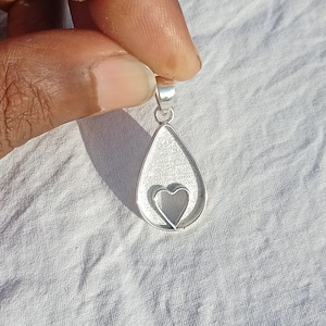 May include: Silver pendant in the shape of a teardrop with a smaller heart cutout inside. The pendant has a textured surface.