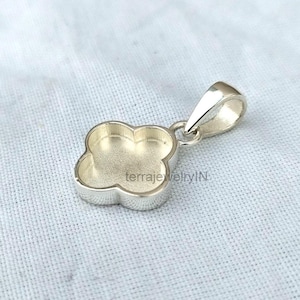 May include: A silver-colored clover-shaped pendant with a loop for a chain. The pendant has a smooth, polished finish and a shallow, concave center. The text "terrajewelryIN" is visible on the pendant.