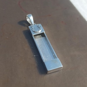 May include: A silver pendant with a rectangular body and a horseshoe design. The pendant has a loop at the top for a chain. The rectangular body has a hollowed-out section. The pendant is made of a shiny, metallic material.