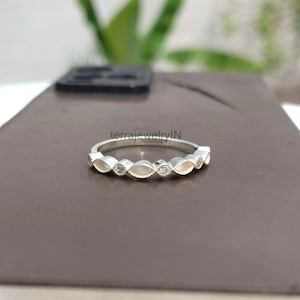May include: A silver ring with a repeating pattern of marquise-shaped links and small, round, clear gemstones. The ring is set on a brown surface with a blurred green background. The text "terrajewelryIN" is visible on the ring.