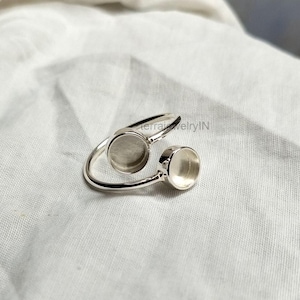 Peut inclure: Une bague réglable argentée avec deux sertissages circulaires. La bague a un design incurvé, avec les sertissages positionnés aux extrémités opposées de la bande. La bague est présentée sur un fond de tissu blanc.