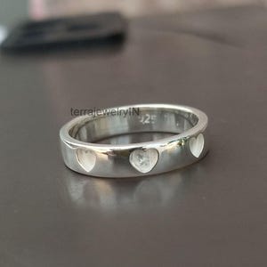 Könnte beinhalten: Ein silberner Ring mit herzförmigen Aussparungen. Der Ring besteht aus poliertem Metall und weist drei herzförmige Aussparungen auf, die gleichmäßig um das Band angeordnet sind. Der Ring wird auf einer dunklen Oberfläche präsentiert. Der Text "terrajewelryIN" und "925" sind sichtbar.