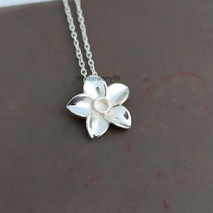 May include: A silver-colored flower pendant necklace. The pendant has five petals and a circular center. The chain is also silver and has a delicate link design. The necklace is displayed on a brown surface.