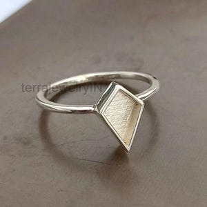 May include: A silver ring with a diamond-shaped setting. The ring's band is thin and circular, leading to the geometric centerpiece. The diamond shape has a textured, light-colored surface, creating a contrast with the polished silver.