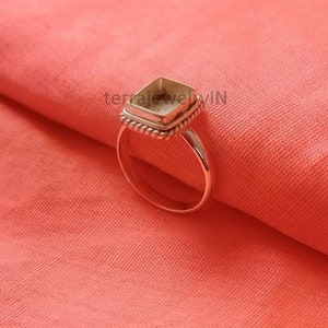 Op de afbeelding: Een zilveren ring met een vierkante zetting, klaar om te worden gevuld met een edelsteen. De ring heeft een touwachtig ontwerp rond de zetting.