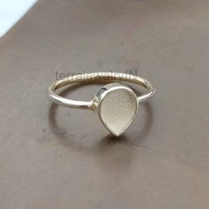 May include: A silver ring with a teardrop-shaped setting. The ring band is thin and polished, leading to the textured, pear-shaped centerpiece. The ring is set against a neutral background.