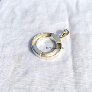 Könnte beinhalten: Ein silber- und goldfarbener Anhänger mit kreisförmigem Design. Der Anhänger hat eine Ringform mit einem goldfarbenen Innenkreis und silberfarbenen Außenseiten. Der Anhänger liegt auf einem weißen Stoff.