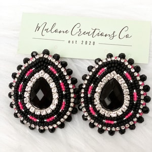 May include: Two black beaded earrings with a teardrop-shaped black centre stone, surrounded by white and pink beads. The earrings have a circular design with a beaded edge. A light green card with the text "Malone Creations Co est 2020" is in the background.