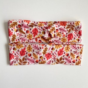 Puede incluir: Un par de diademas de tela blanca con un estampado de oto&ntilde;o de hojas rojas, naranjas y amarillas, bellotas y bayas.