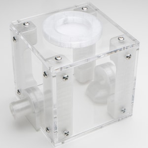 May include: A clear, cube-shaped container with a circular opening on top and a cylindrical port on the side. The container is constructed of transparent acrylic panels held together with small silver screws. Interior components are visible within the cube.