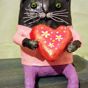 May include: A black cat figurine wearing a pink shirt and purple pants holds a red heart with white flowers painted on it.