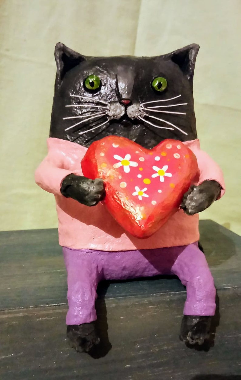 May include: A black cat figurine wearing a pink shirt and purple pants holds a red heart with white flowers painted on it.