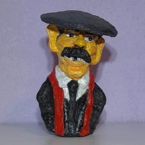 Pode incluir: Uma figurinha feita à mão de uma pessoa usando um boné preto, um terno preto e uma echarpe vermelha. A figurinha tem o rosto amarelo, bigode preto e dentes brancos.