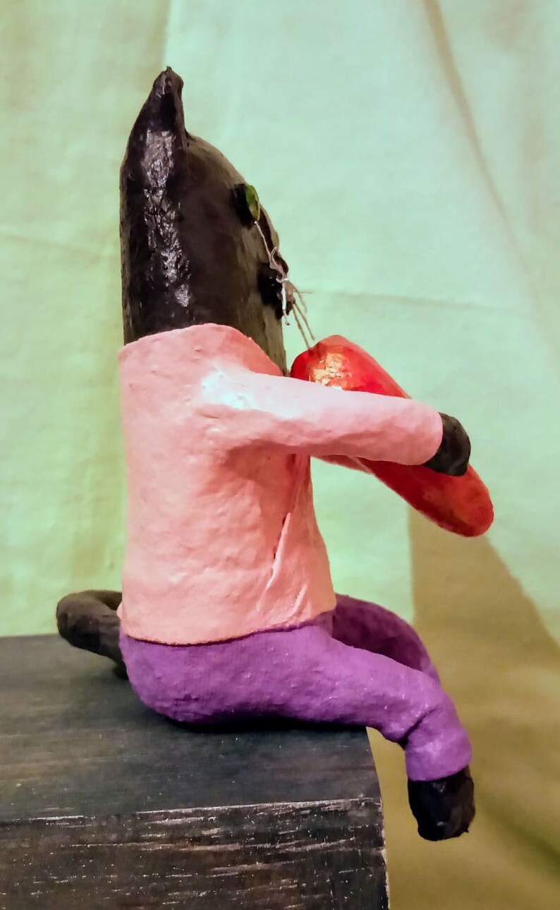 May include: A black cat figurine wearing a pink shirt and purple pants, holding a red heart in its paws.