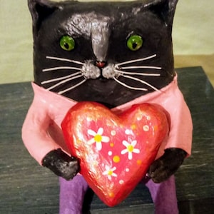 May include: A black cat figurine wearing a pink shirt and holding a red heart with white flowers. The heart is decorated with orange dots.